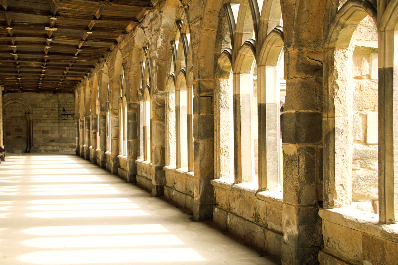 Dimly lit stone corridor in Durham Cathedral, with Gothic arches and sunlight streaming through windows, creating patterns on the floor.