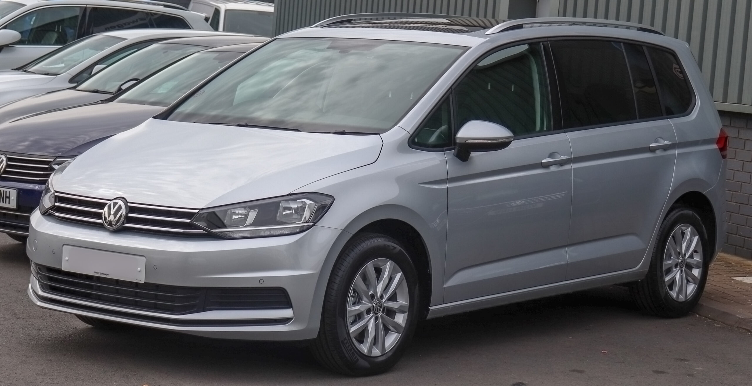 A silver Volkswagen car is parked among other vehicles in a lot, near a building with vertical metal siding.