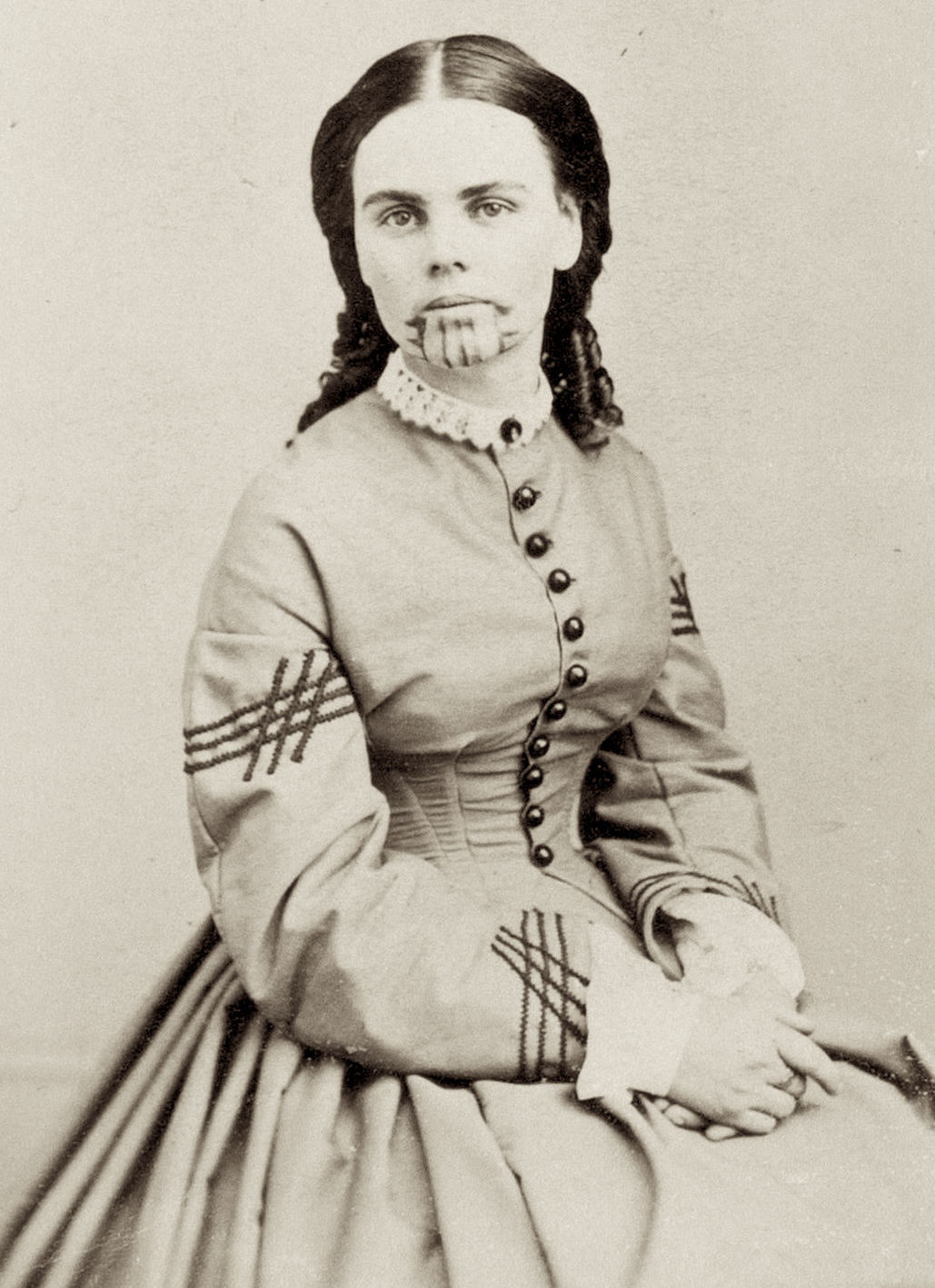 A person in a 19th-century dress sits with a distinctive chin tattoo, looking directly at the camera, showcasing historical fashion and cultural markings.
