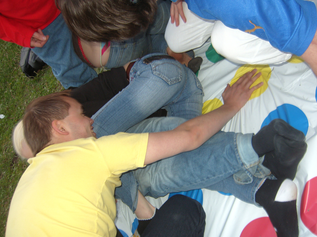 People playing Twister on grass, intertwined in colorful positions. Participants wear casual clothing, creating a fun and energetic outdoor scene.