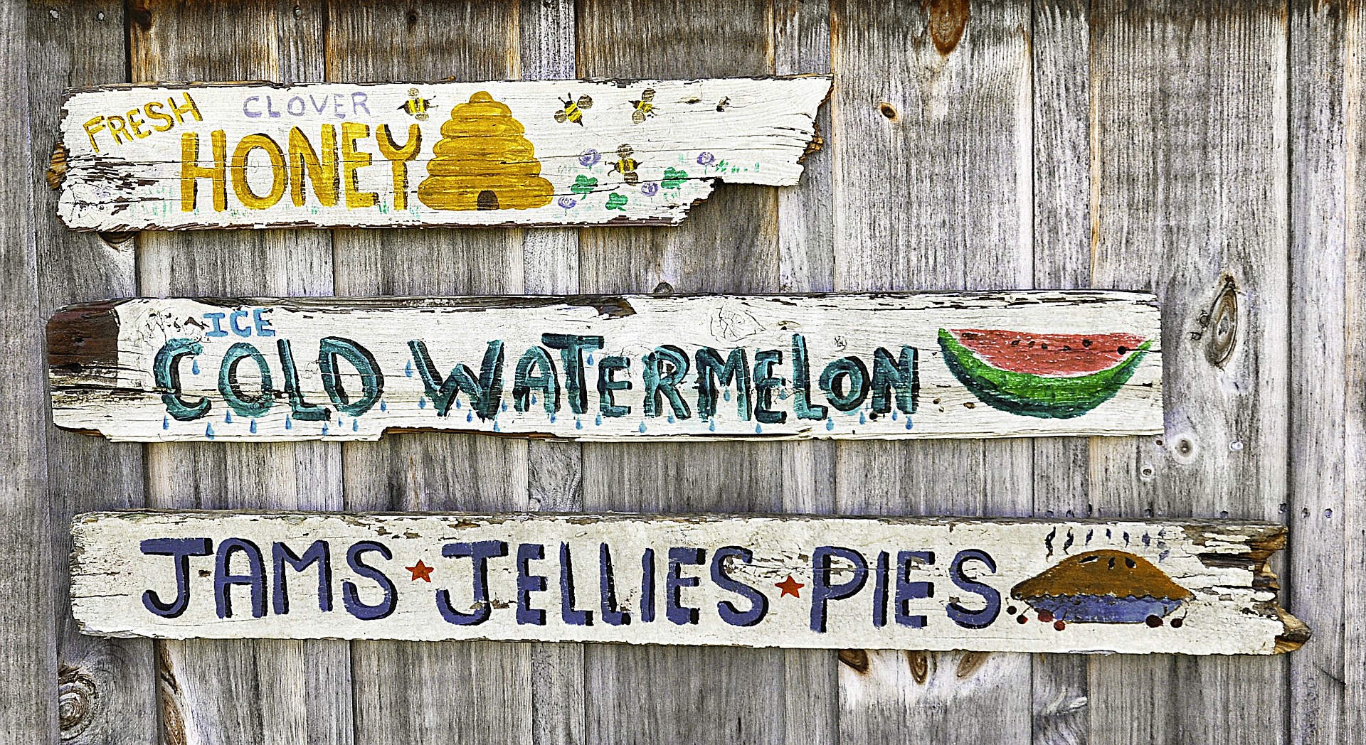 Three rustic wooden signs promote fresh clover honey, cold watermelon, and jams, jellies, and pies, displayed against a weathered wooden fence.