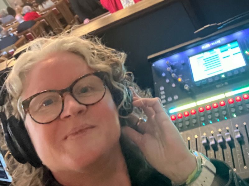 A person with headphones stands by a mixing console in a busy indoor setting, appearing engaged and focused on audio work.