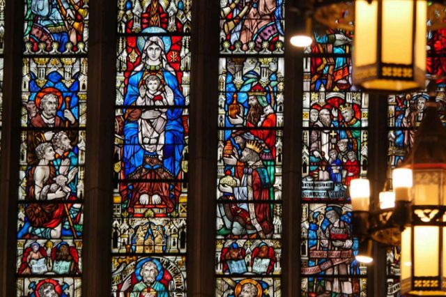 Vibrant stained glass windows depicting religious scenes, with intricate designs and figures, in a historical church setting illuminated by ornate hanging lamps.