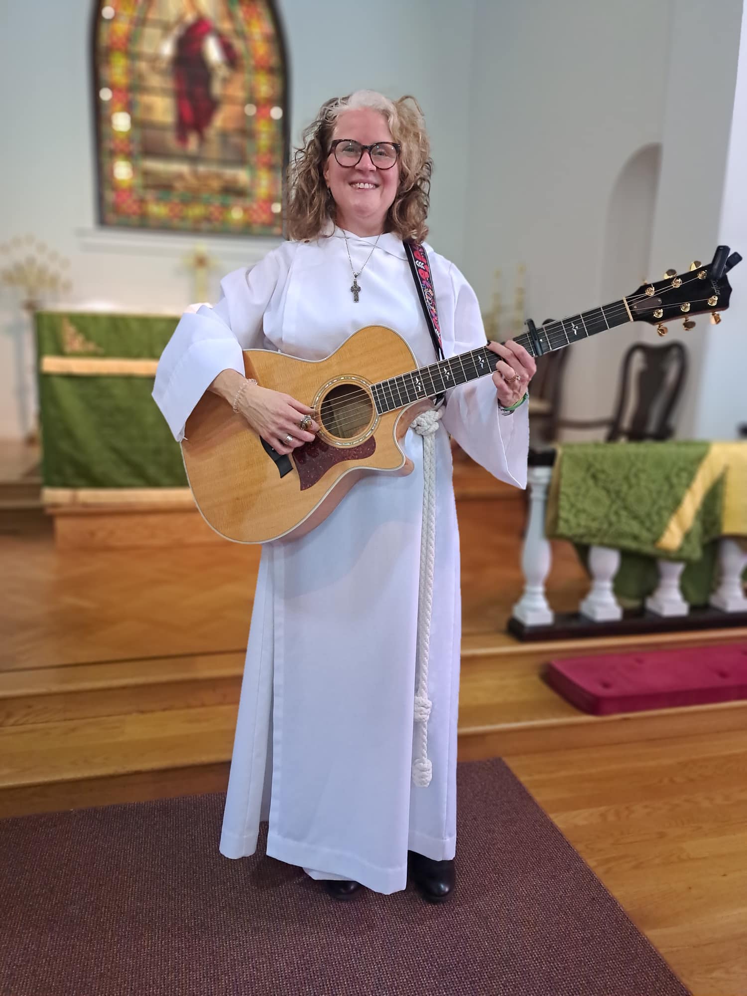 A person in a white robe stands in a church, playing a guitar with a stained glass window in the background.