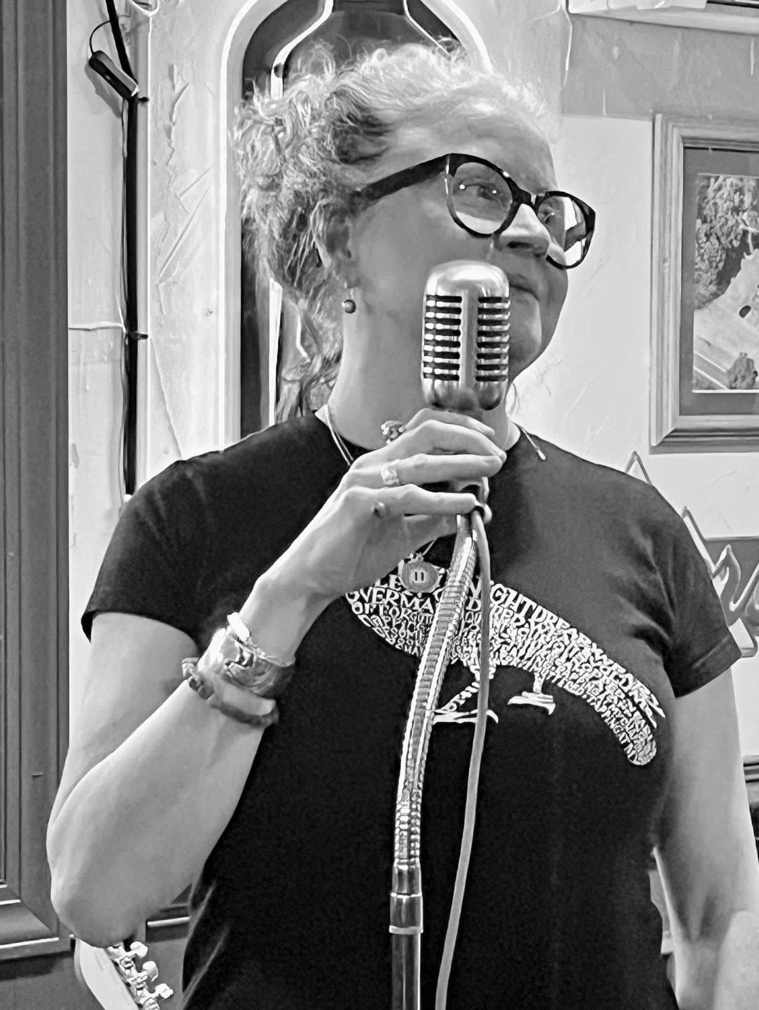 A person wearing glasses holds a vintage microphone in a room with framed artwork and decorative elements.